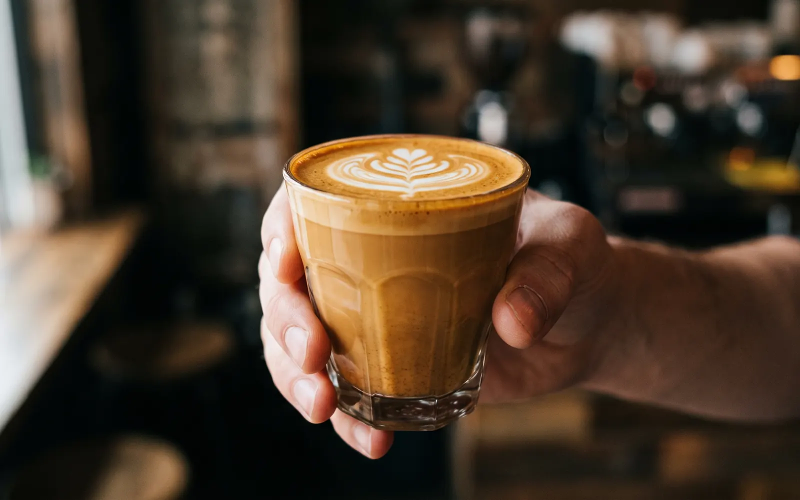 A hand holding a cortado in a glass in a dimly lit cafe