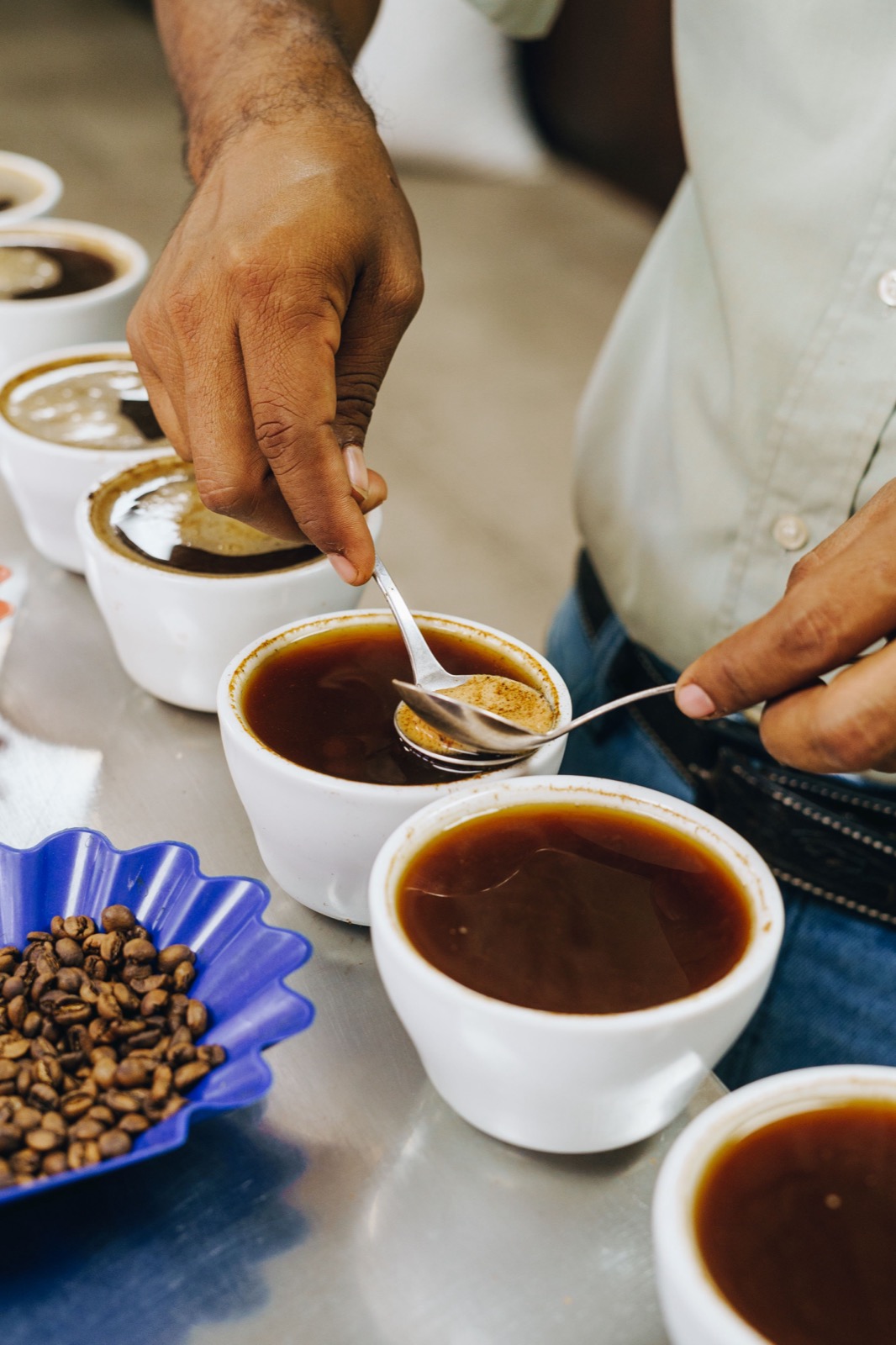 Close-up of a cupping spoon lifting coffee from a sample cup beside a tray of green beans